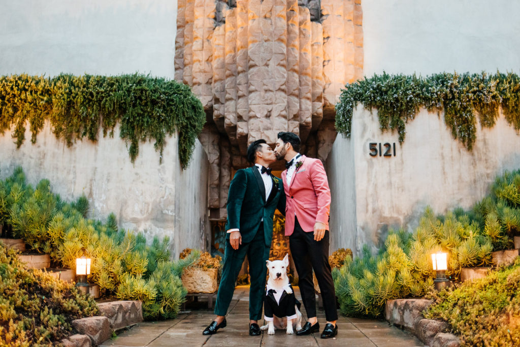 Two grooms kissing in front of the historic Sowden House with their rescue dog