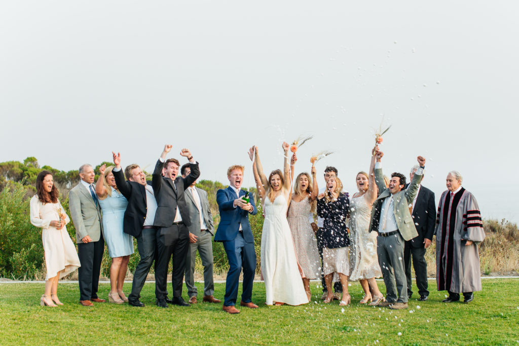 wedding party and family cheer as groom pops a bottle of champagne