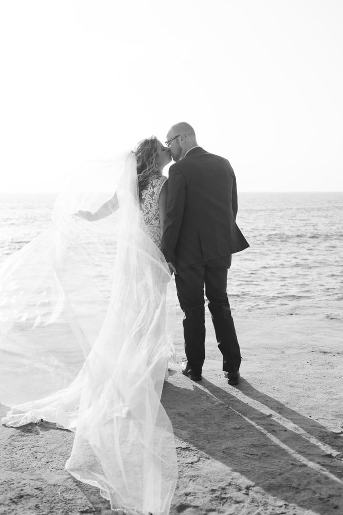 bride and groom kiss at sunset with the veil flowing in the wind