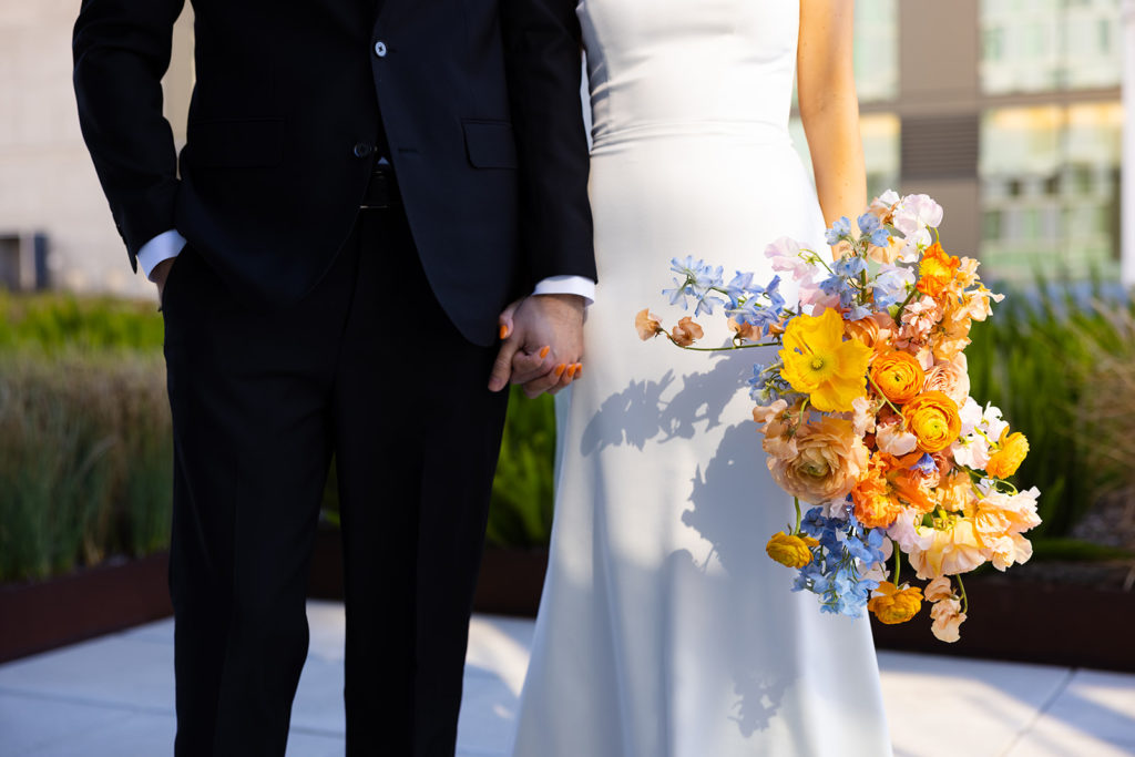 bride and groom hold hands while she grips her colorful and whimsical wedding bouquet