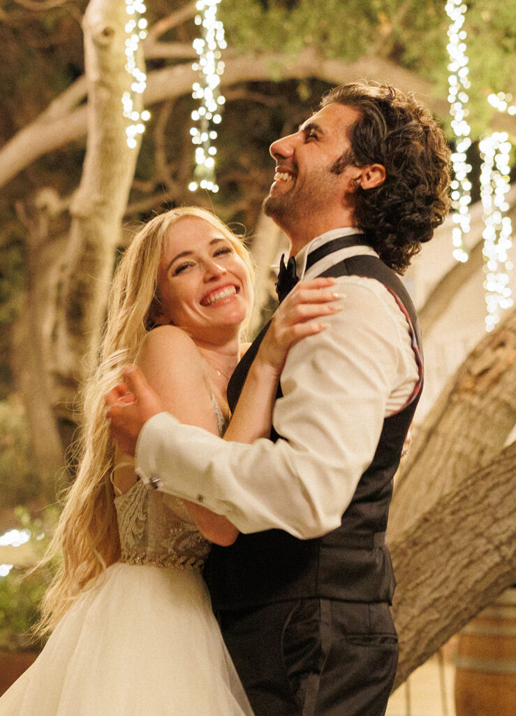bride and groom giggle as they hold each other in a close embrace under twinkle lights