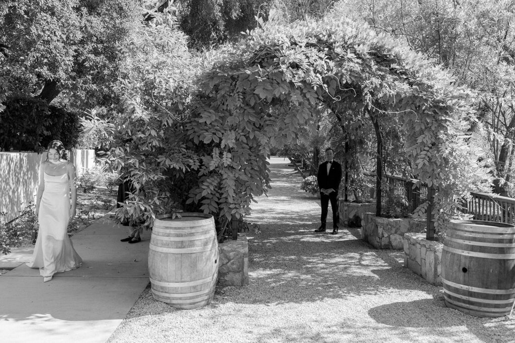 wide view of bride walking to her first look with the groom under the vine-covered pathway at Calamigos Ranch