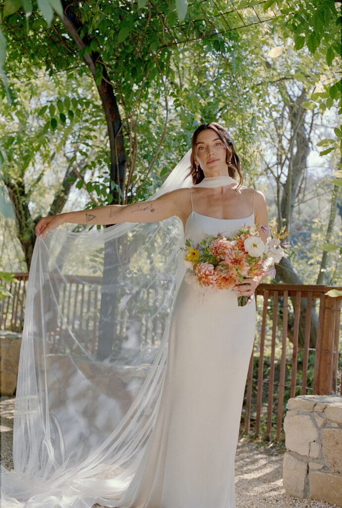bride in modern satin wedding dress and white neck scarf holds her colorful wildflower bouquet and fluffs her veil under the trees at Calamigos Ranch