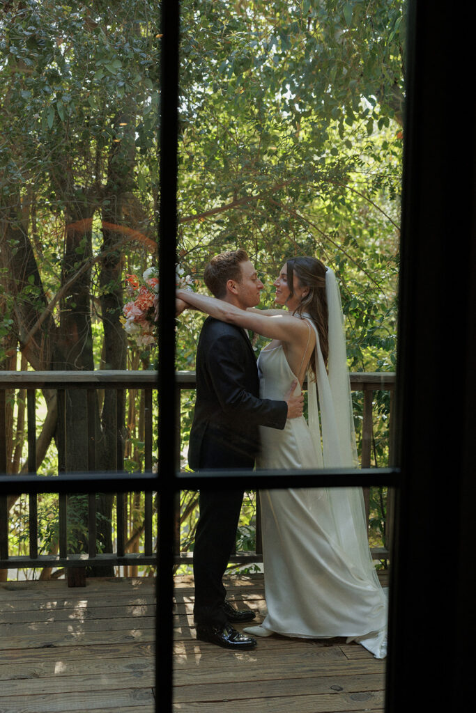 view through the window of bride and groom embracing on the wood deck underneath the trees at Birchwood Room