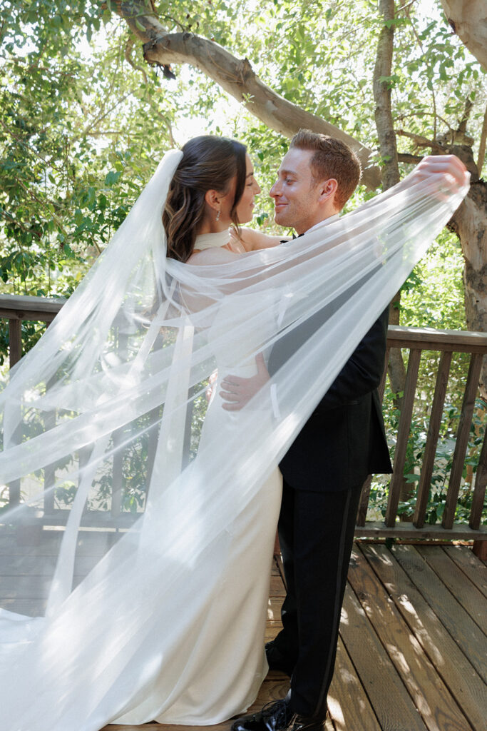 bride sweeps her veil around her and the groom for a romantic embrace on the wood deck under the trees at Birchwood Room in Calamigos Ranch